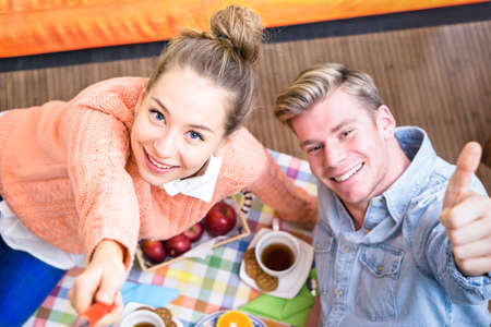 Cheerful couple taking selfie at breakfast lying on the living room floor - Happy young friends share fun moments together using mobile phone stick at home - Concept of daily healthy life togetherの写真素材