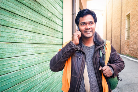 Young indian travel man making phone call walking on the street holding bag at winter time - Handsome bangladeshi guy looking up using mobile smiling outdoors  - Communication concept with copy spaceの写真素材