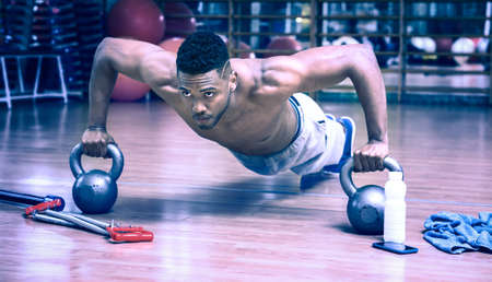 African american man doing push up workout in dark gym using kettlebell - Black gritty face guy working on chest muscles exercise at fitness center - Bluish vintage filter look with dim light effectの写真素材