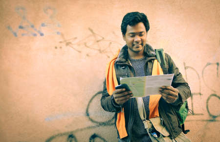 Indian tourist man looking map leaning at grunge urban wall background - Travel portrait of young bangladeshi guy searching street on guide casual fall wear - Vintage warm filter vignetting effectの写真素材