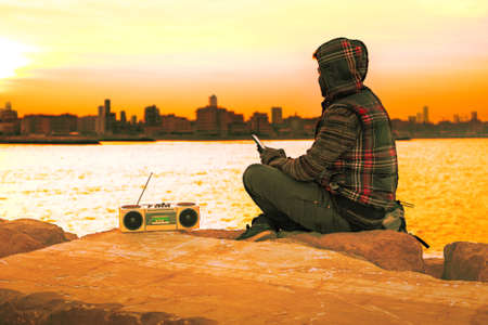 Pensive african american man silhouette sitting by the ocean looking at cityscape - Hooded guy listening music holding phone with melancholic attitude - Concept of dreams and hopes with golden filterの写真素材