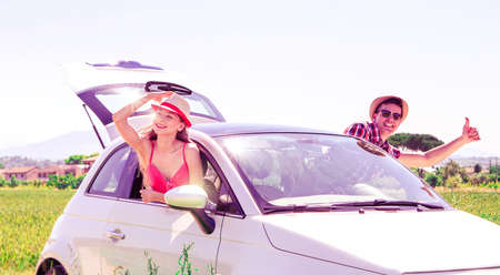 Car happy couple popping body out the window - Cheerful young teenagers road trip vacation with thumb up and positive attitude - Summer journey moments of fun driving new automobile at countrysideの写真素材