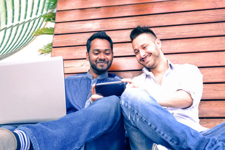 Young interracial couple of happy men using phone sitting on urban wall background - Happy indian...