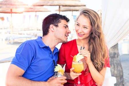 Happy couple drinking cocktails in beach bar at sunset - Young lovers smiling on  vacation dating moments holding drinks with male whispering at beautiful blonde female - Concept of summer love storyの写真素材