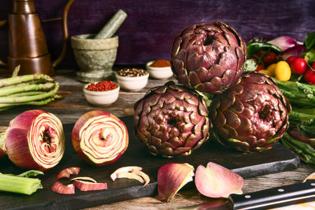 Fresh roman artichokes on cutting board with spices and cooking tools  in dark background - Spring vegetables on rustic kitchen table front angle view with morning light and shadowsの写真素材