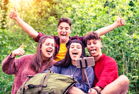Group of young friends taking selfie with stick on green forest background - Teenagers having fun using mobile phone technology for self photo on camping excursion day - Soft vintage filter lookの写真素材