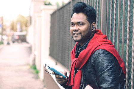 Young indian guy with headsets listening music holding mobile phone outdoors - Handsome bangladeshi...