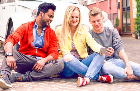 Happy multiracial friends having fun taking selfie on road trip - Multicultural teenagers using mobile phone camera sitting outdoor near vintage car - Concept of friendship travel and new technologiesの写真素材