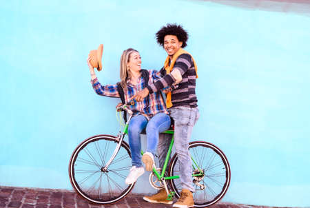 Happy interracial couple riding sport bike on blue sky wall background - Playful friends on bicycle tour having fun moments together - Concept of healthy lifestyle and travel in tilted compositionの写真素材