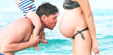 Dad kissing pregnant woman belly on the beach giving at child a piggyback ride - Happy family having fun moments by the ocean on summer vacation day - Concept of love and motherhood emotions - Imageの写真素材