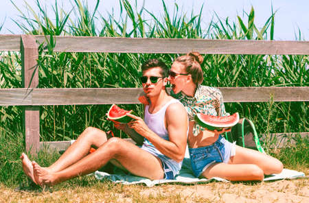 Funny moment for friends eating watermelon at picnic on the beach - Woman teasing moody man by biting his ear - Young couple on the summer in playful/litigious attitude - Vintage filter look - Imageの写真素材