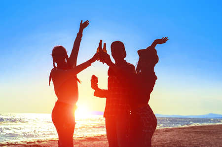 Silhouette of happy friends dance at beach party on sunrise  light - Teenagers cheers with beer bottles having fun  moment on summer holiday - Concept of teenage friendship - Soft bluish filter imageの写真素材