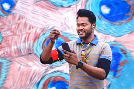 Young indian man holding mobile phone inserting earphone plug at device - Happy asian guy using smartphone headset outdoor in casual sport look - Concept of modern communication technology - Imageの写真素材