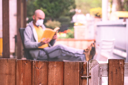 Man with face mask reading book in courtyard main focus on the fence - Concept of social isolation...