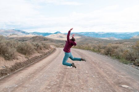 Young caucasian girl woman jumping on the dirt road in rural BC, dry grass covered hills. Enjoying the freedom off road trip adventure.の写真素材