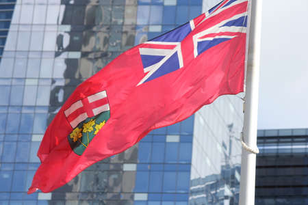 Flag of the Province of Ontario blowing in the breeze against a blue sky, in front of a building of glass windows.の写真素材