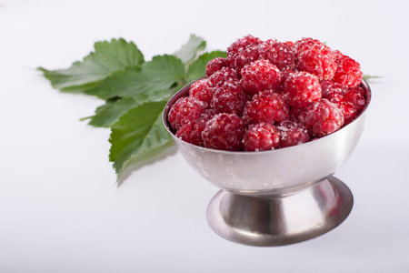 red raspberies in metal cup with leafs behind on white background