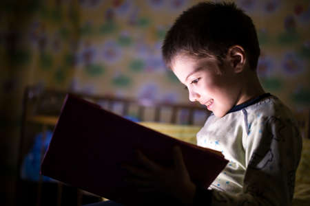 little boy in a dark room, reading a book with interestの写真素材