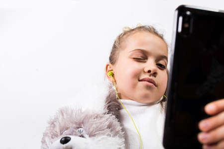 Little girl blogger with earphones looking to smartphone hugging plush toy on a white backgroundの写真素材