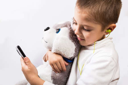 Phone addicted little boy with plush toy and with earphones on white backgroundの写真素材