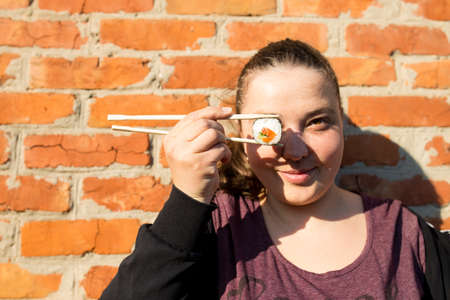Happy girl holding one sushi roll with Chinese chopsticks on a brick wall background.の写真素材