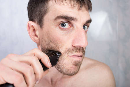 Young man shaving face with electric trimmer, look close to camera with funny face.の写真素材