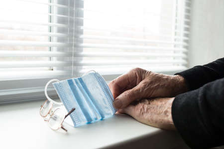 Old human hands holding medical mask next to the window, on self-isolation.の写真素材