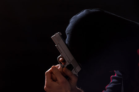 Man with hood on holding gun and praying on black background.