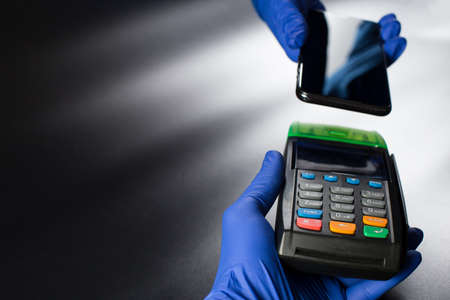 Contactless payment operation, hands in blue gloves holding contactless terminal and smartphone on other hands, on black background with copy space.の写真素材