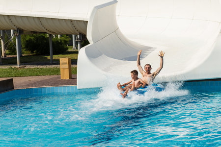 Boy and dad sliding from water hill in water park, durring summer outside.