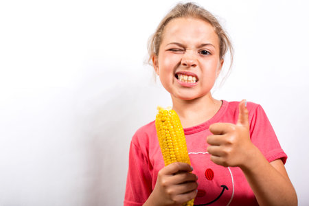 Girl kid holding sweet corn in hand and showing thumb up on a white with copy space.の写真素材