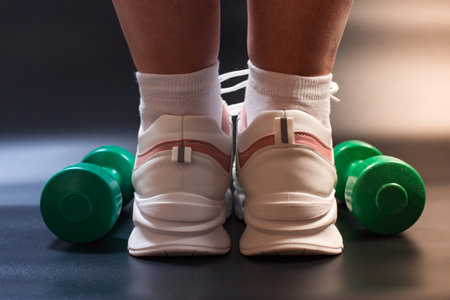 Female foots in white sneakers with green dumbbells close to, close up on black background.