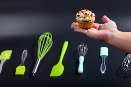 Female hand with cup cake and tools of pastry chef on black background.
