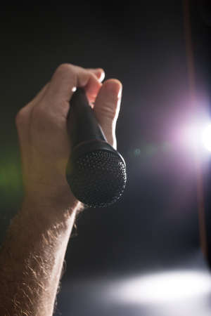 A microphone in the hands of a rock artist clse up with spotlights on a stage, on black background.の写真素材