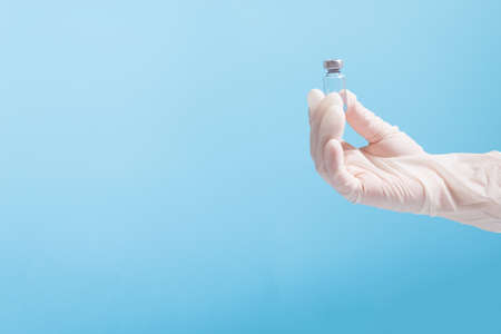 Hand in white glove holding vaccine dose in glass little container, on blue background with copy space.の写真素材
