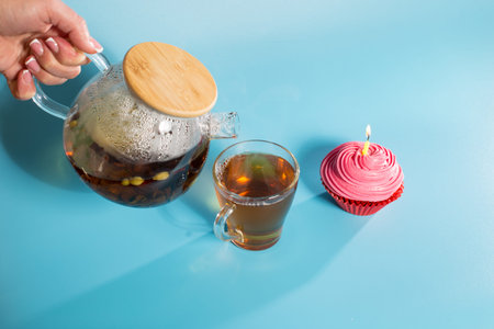 A hand holds a teapot with tea leaves and pours into a mug, on a blue background with birthday...