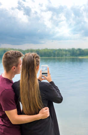 A couple of young man and a girl taking pictures on a smartphone on the lake. Travel and entertainment concept.の写真素材