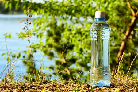 Water in a PET bottle on the lake shore against the background of green tree branches. Environmental problems in the use of natural resources.の写真素材