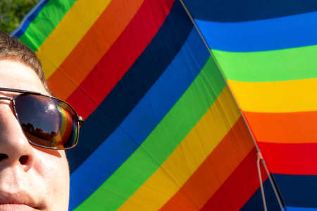 A young man in glasses stands with a rainbow umbrella, a symbolic color logo icon for equal rights in love and marriage social equalityの写真素材