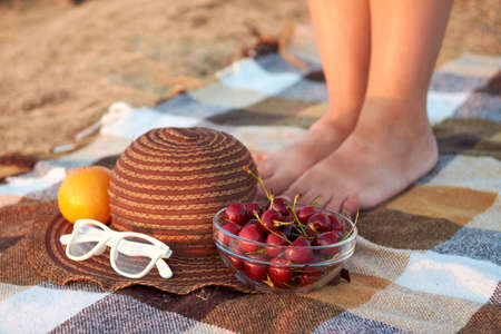 On the checkered bedspread is a hat, glasses, a plate with a cherry on the background of women's legs. Picnic in nature.の写真素材