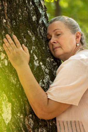 an elderly woman embraces a tree trunk, the concept of connecting with nature, promoting healthの写真素材
