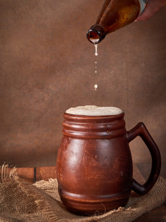 Beer clay brown mug with beer on a wooden table on a dark background. A stream of light beer pours into a glassの写真素材