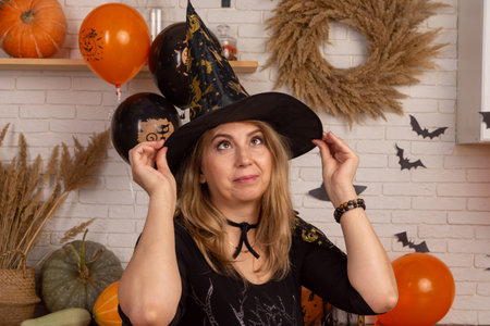 A beautiful woman in a black dress puts on a witch hat on a background decorated for Halloween....