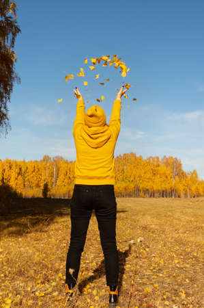 A woman standing with her back throws yellow autumn leaves into the air in a park on an autumn sunny day. Portrait of a woman in yellow clothes playing with autumn leaves in the forestの写真素材