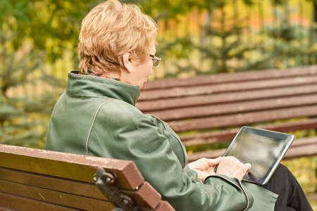 an elderly woman with a tablet computer is sitting on a bench in an autumn park. The concept of technologies for remote communication of elderly people with their relativesの写真素材