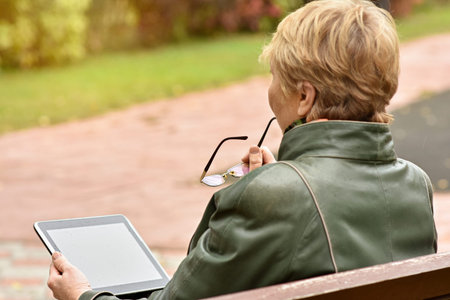an elderly woman with a tablet computer is sitting on a bench in an autumn park. The concept of technologies for remote communication of elderly people with their relativesの写真素材