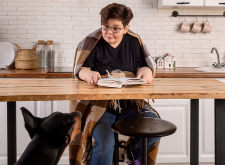 A young boy wearing glasses is sitting at a kitchen table reading a book. The boy is wrapped in a blanket. The dog is sitting beside the table on the floor looking up at the boy.の写真素材