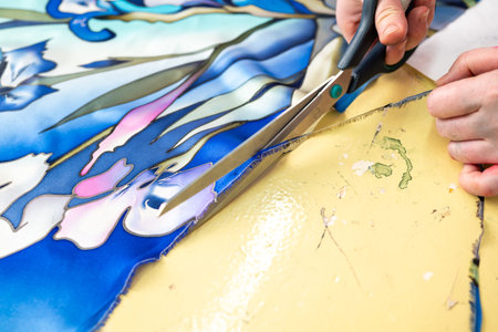 Close-up of a person s hands cutting a piece of blue and green floral fabric with a pair of...