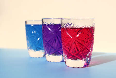 Three crystal glasses with bright neon drinks. Blue, beige background. Food coloring, paint mixing. Purple, pink, turquoise water. Sunlight, glare, highlights on faceted dishesの写真素材