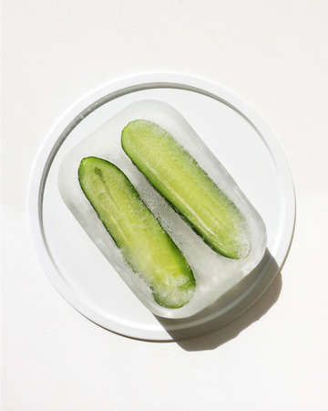 White plate with green cucumber in rectangular ice. Hard light, flat lay. Frozen organic food. Preservation of summer vitamins. Concept of shock freezing of vegetable. Vertical posterの写真素材